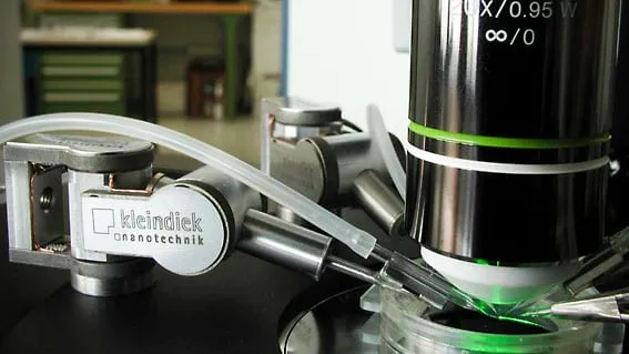 Two MM3A-LS manipulators positioning glass pipettes at the objective of an upright light microscope, illuminated by a green fluorescence ring.